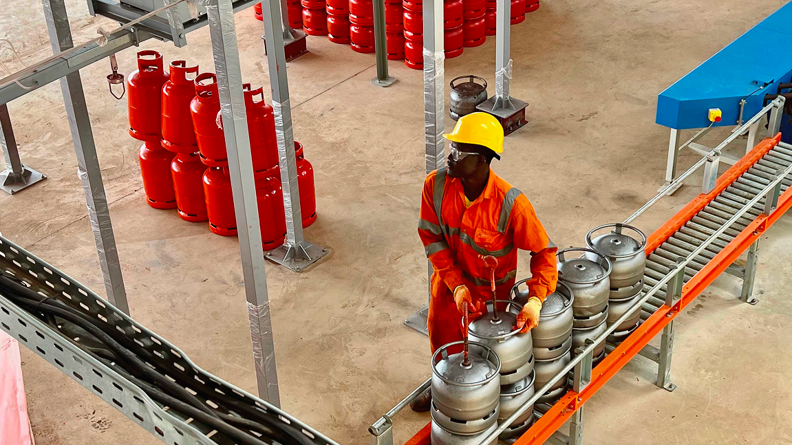 Worker in orange uniform moving gas cylinders on a factory conveyor line.