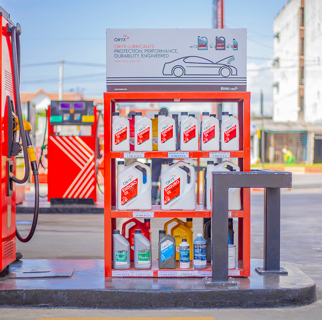 Shelves of motor oil and car fluids at a gas station, with pumps and buildings in background.
