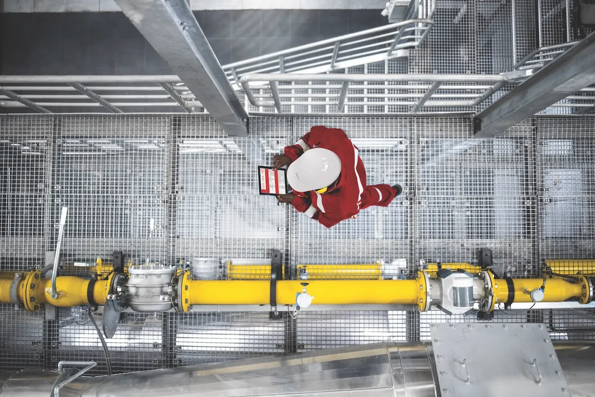 Worker in red protective gear and helmet walking on a metal platform near a yellow pipe.