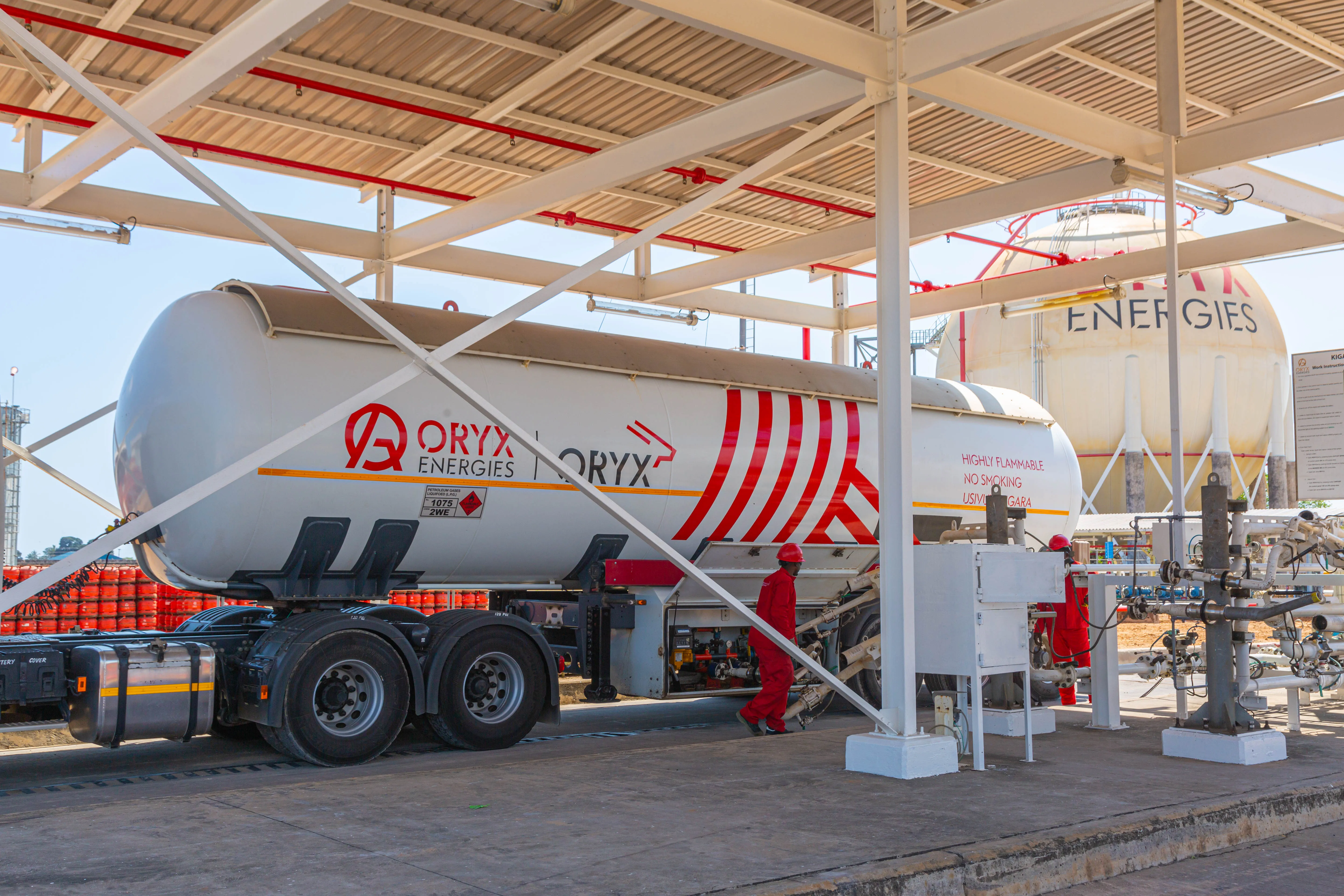 A large fuel tanker truck at an energy facility with equipment and a worker in red uniform.