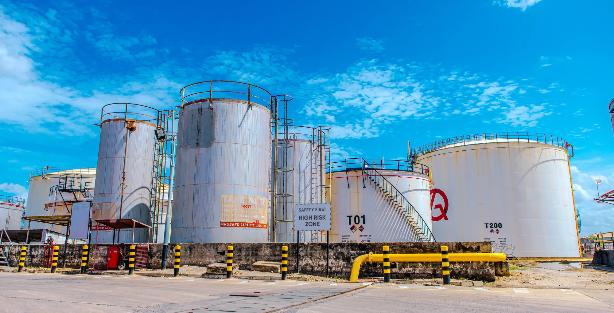 Large industrial fuel storage tanks under a bright blue sky with safety barriers in front.