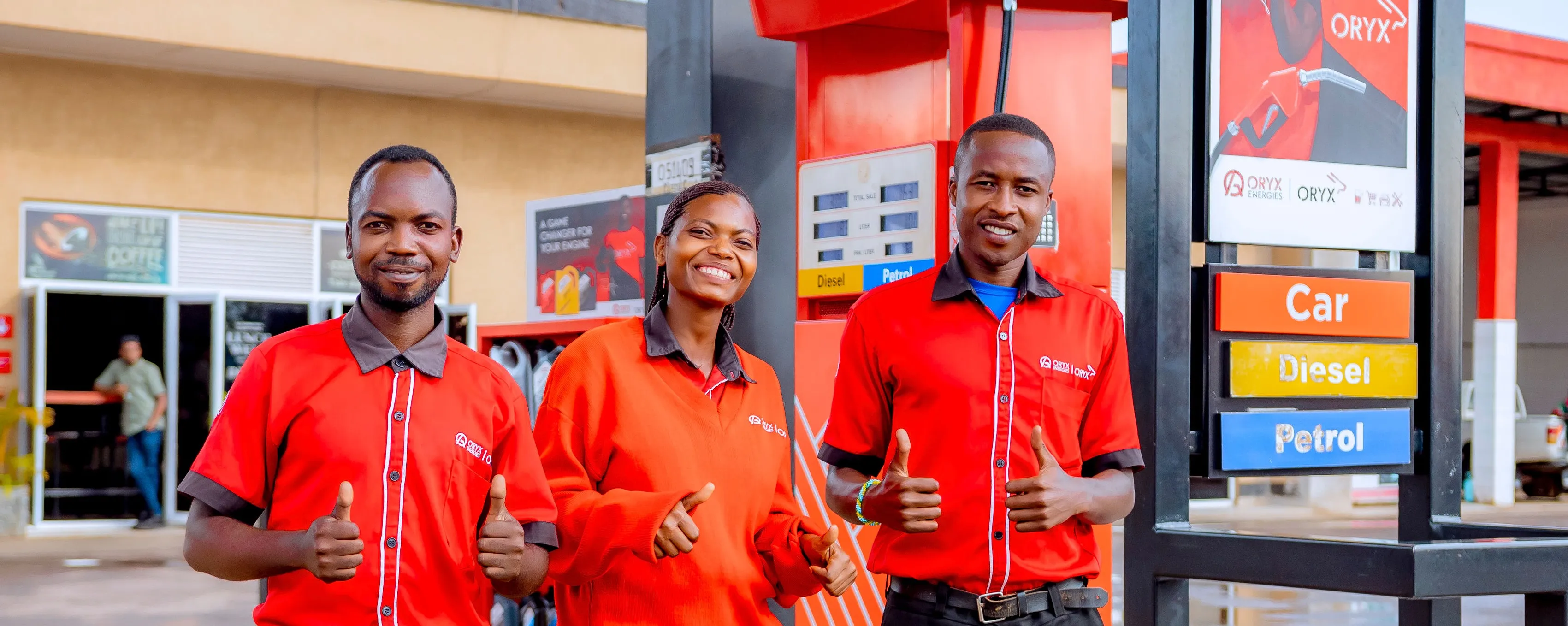 Three smiling gas station attendants in red uniforms give thumbs up at a fuel pump.