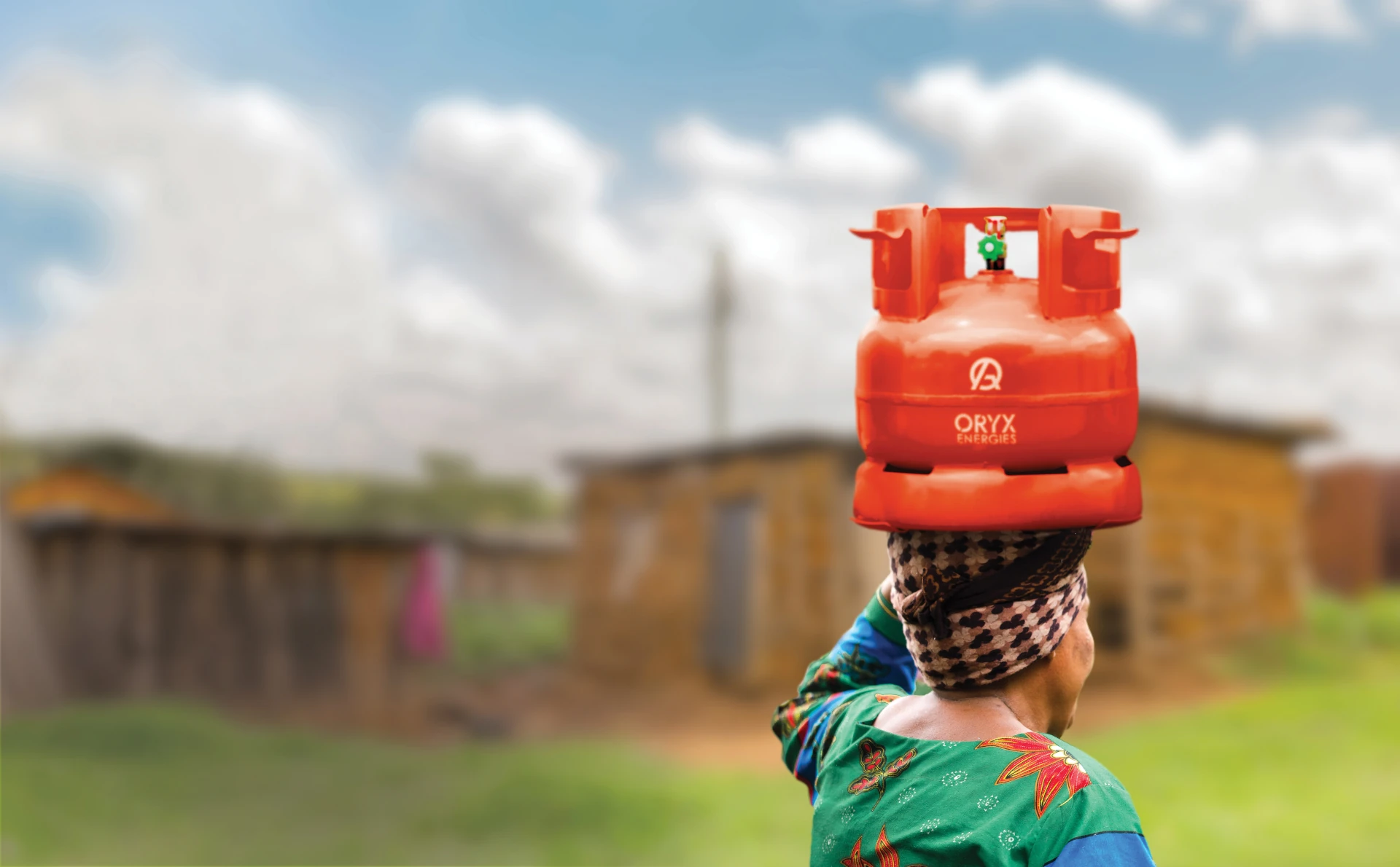 Woman carrying a red gas cylinder on her head in a rural area with blurred houses.