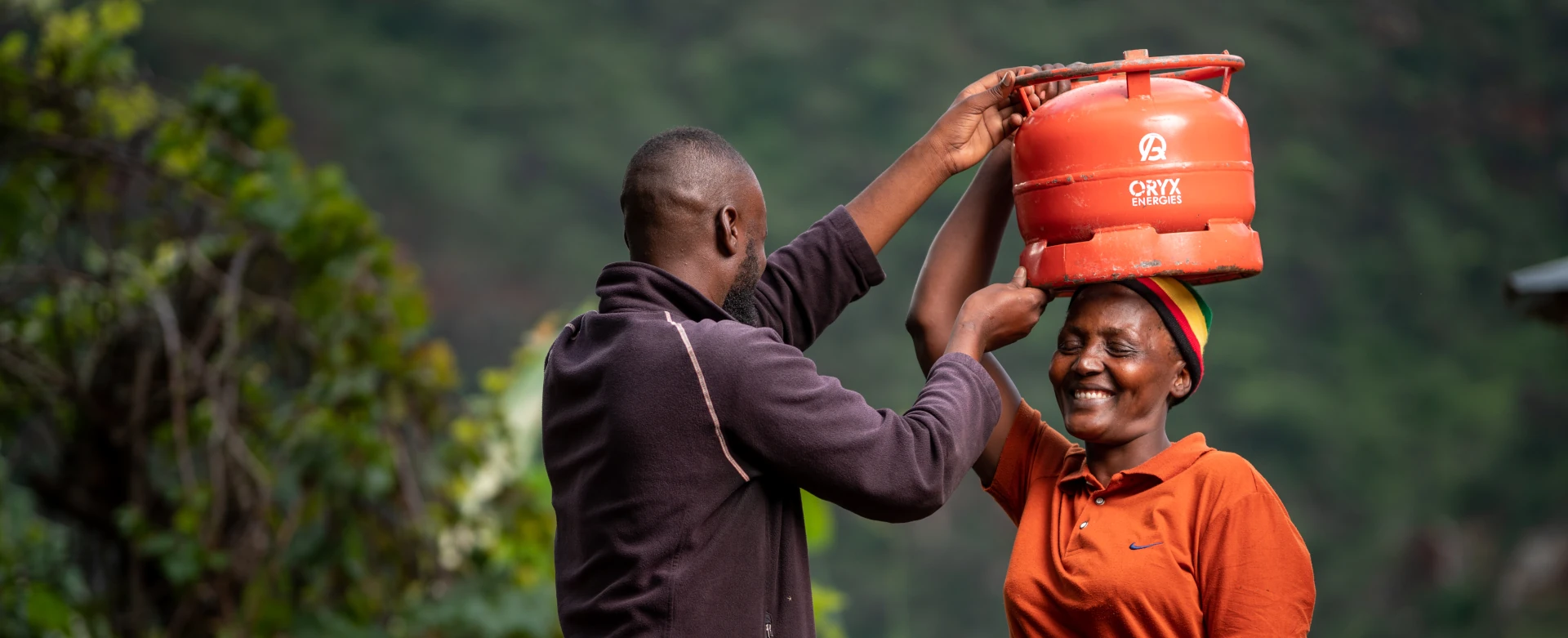 Man helping woman balance a red gas cylinder on her head outdoors, both smiling.