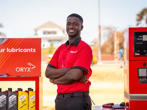 Smiling gas station attendant in red uniform standing by fuel pump and lubricants display.